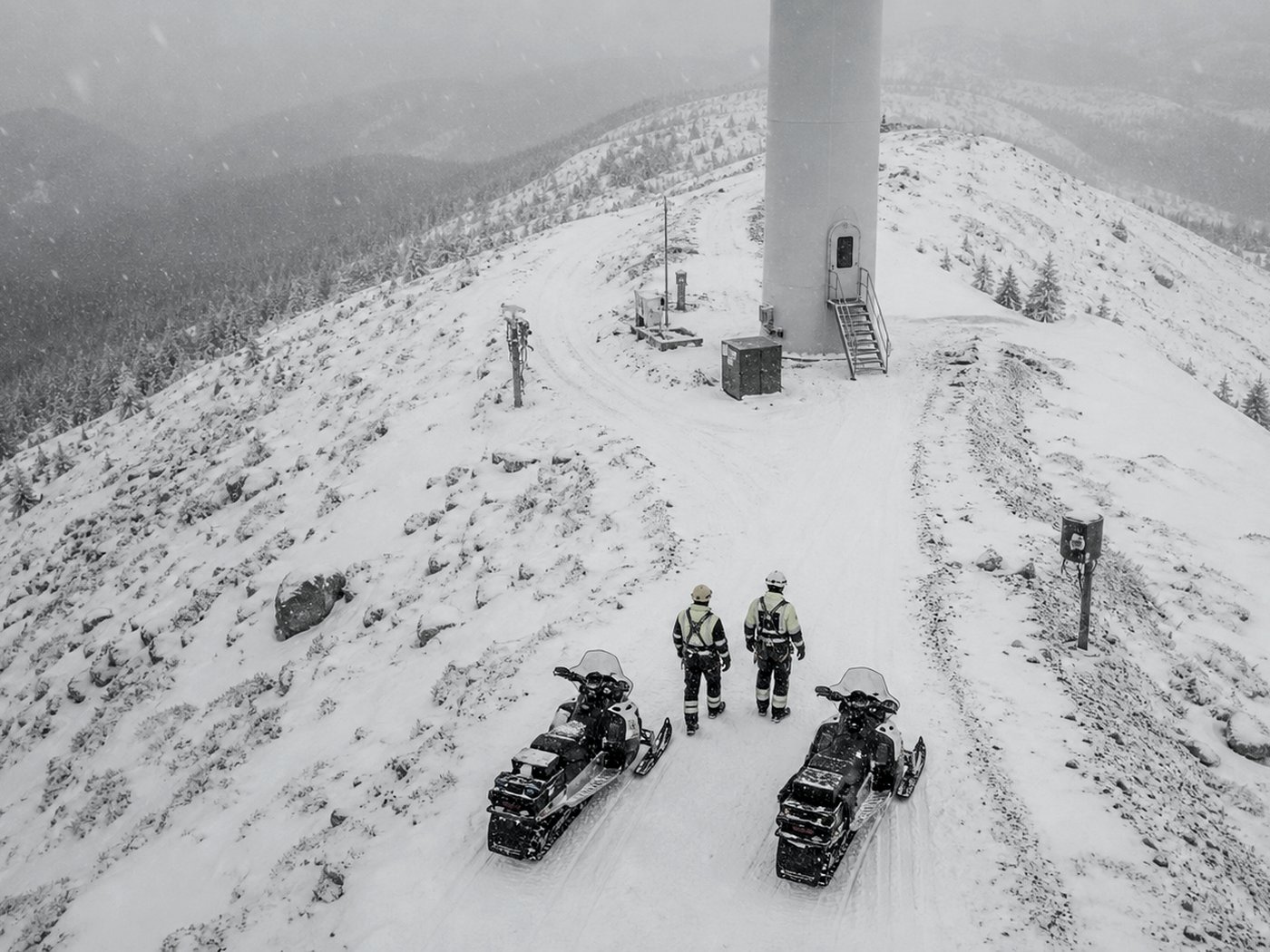 Aerial view of two crew snowmobiles parked at the base of a wind turbine tower on a snow-covered ridge, with two crew members in harnesses standing nearby.